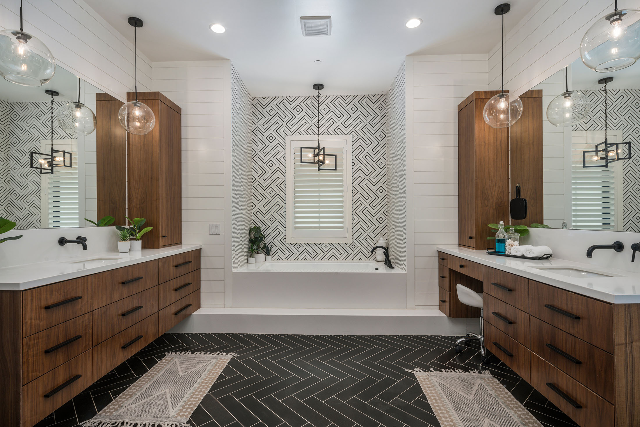 Modern Double Vanity Bathroom with Geometric Tile Design – A stylish bathroom featuring dual wooden vanities, geometric patterned backsplash, pendant glass lighting, and a built-in bathtub. The black herringbone tile flooring adds contrast, creating a luxurious and contemporary atmosphere.
