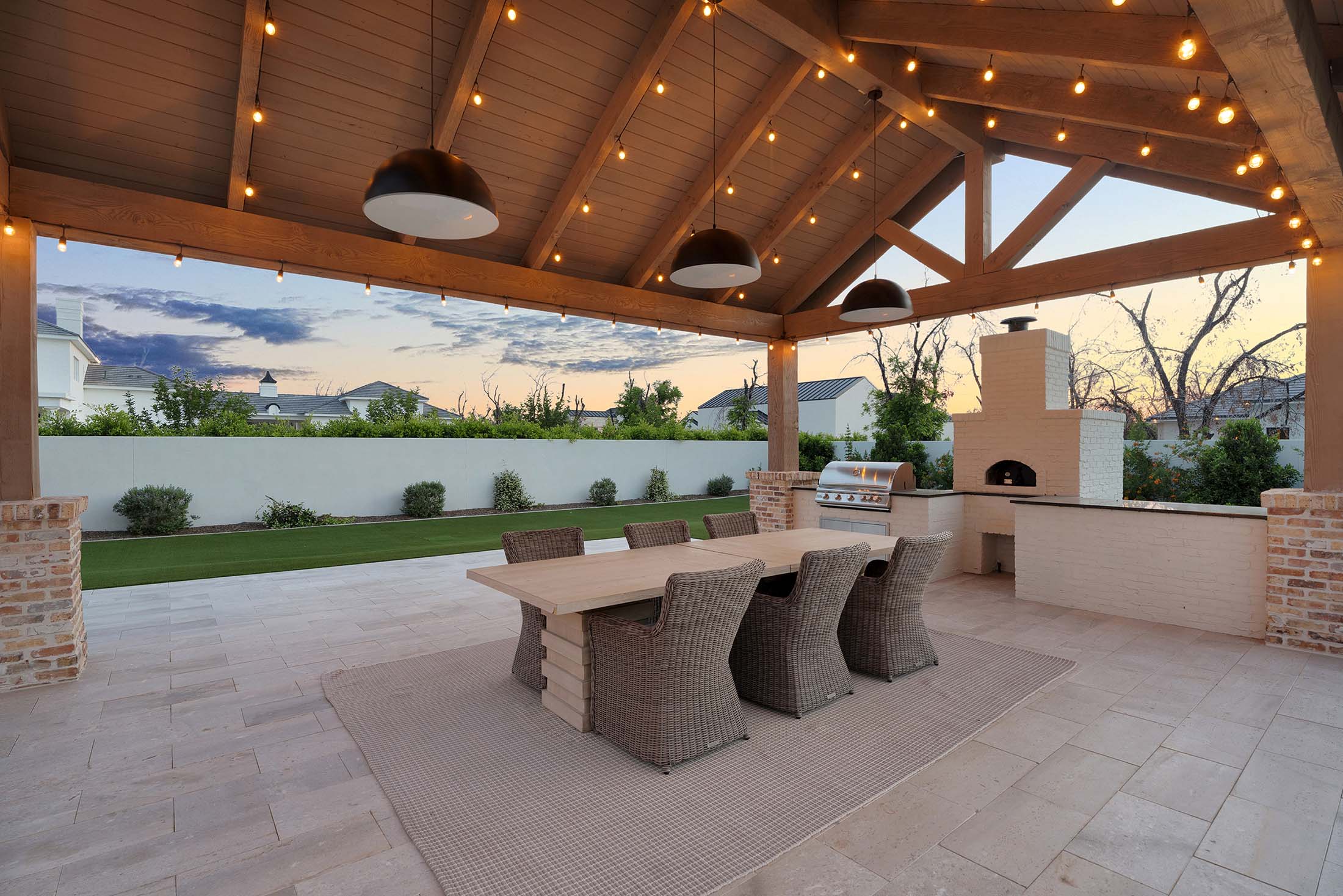 Expansive covered outdoor living space with a built-in brick pizza oven, stainless steel grill, and a rustic wood dining table surrounded by woven wicker chairs. The vaulted wood beam ceiling and black pendant lighting create a cozy yet refined entertaining space.