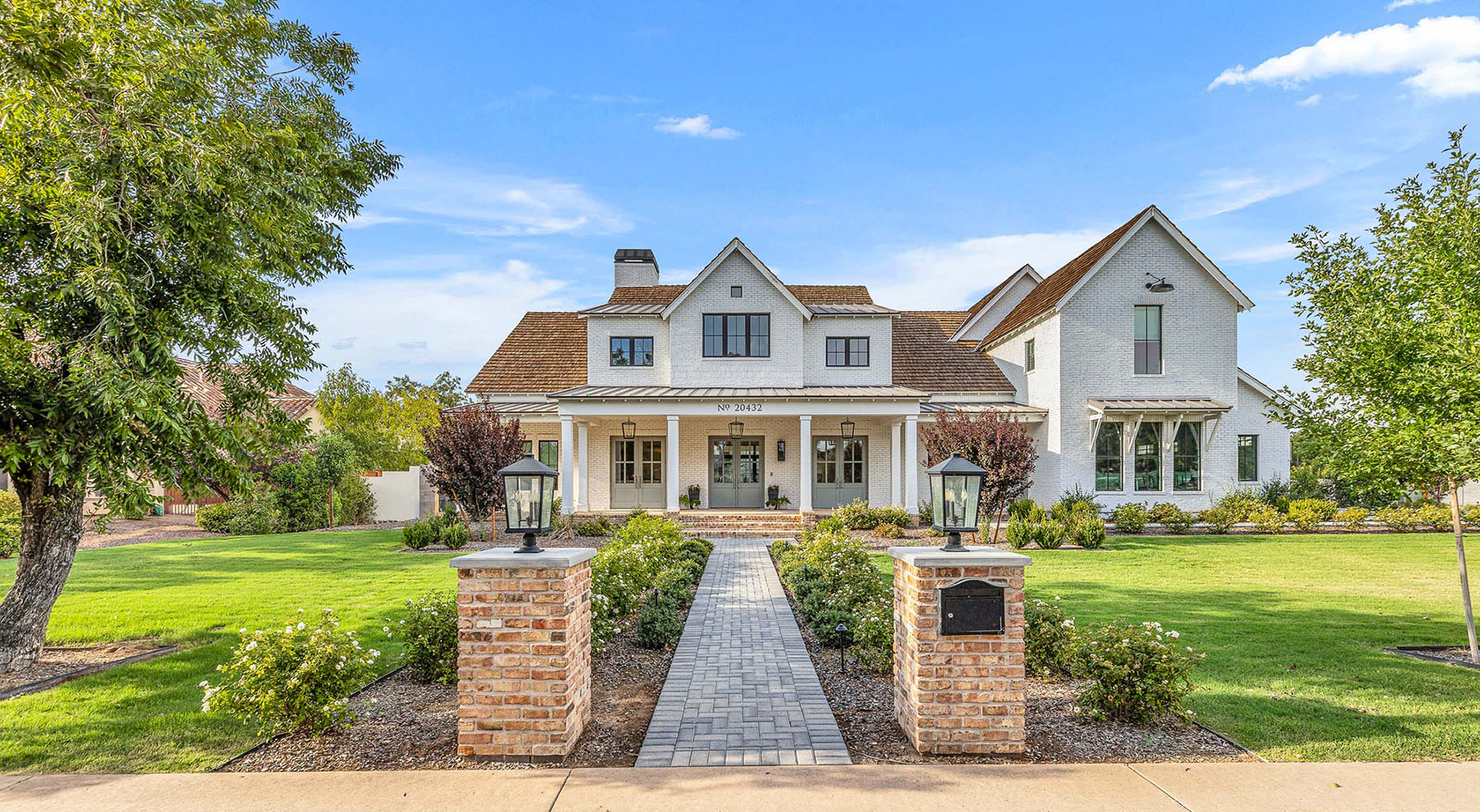 Luxury custom white brick home with a welcoming front porch, elegant landscaping, and timeless architectural details. Featuring classic wood accents, large glass-paneled doors, and lush greenery, this high-end residence embodies refined craftsmanship and sophisticated living.